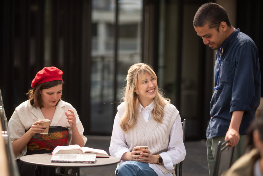 Three students around an outdoor table reading and smiling