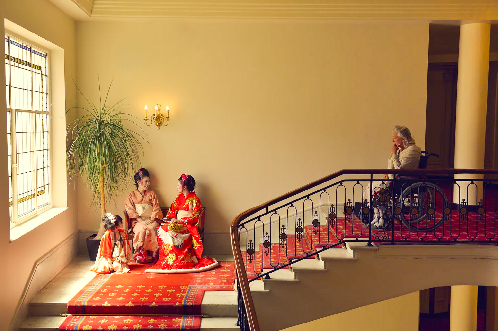 Hallucinations, an artwork by Amaia Salazar. An old woman in a wheelchair looks on at two women and a child in traditional Japanese dress sat on a staircase.