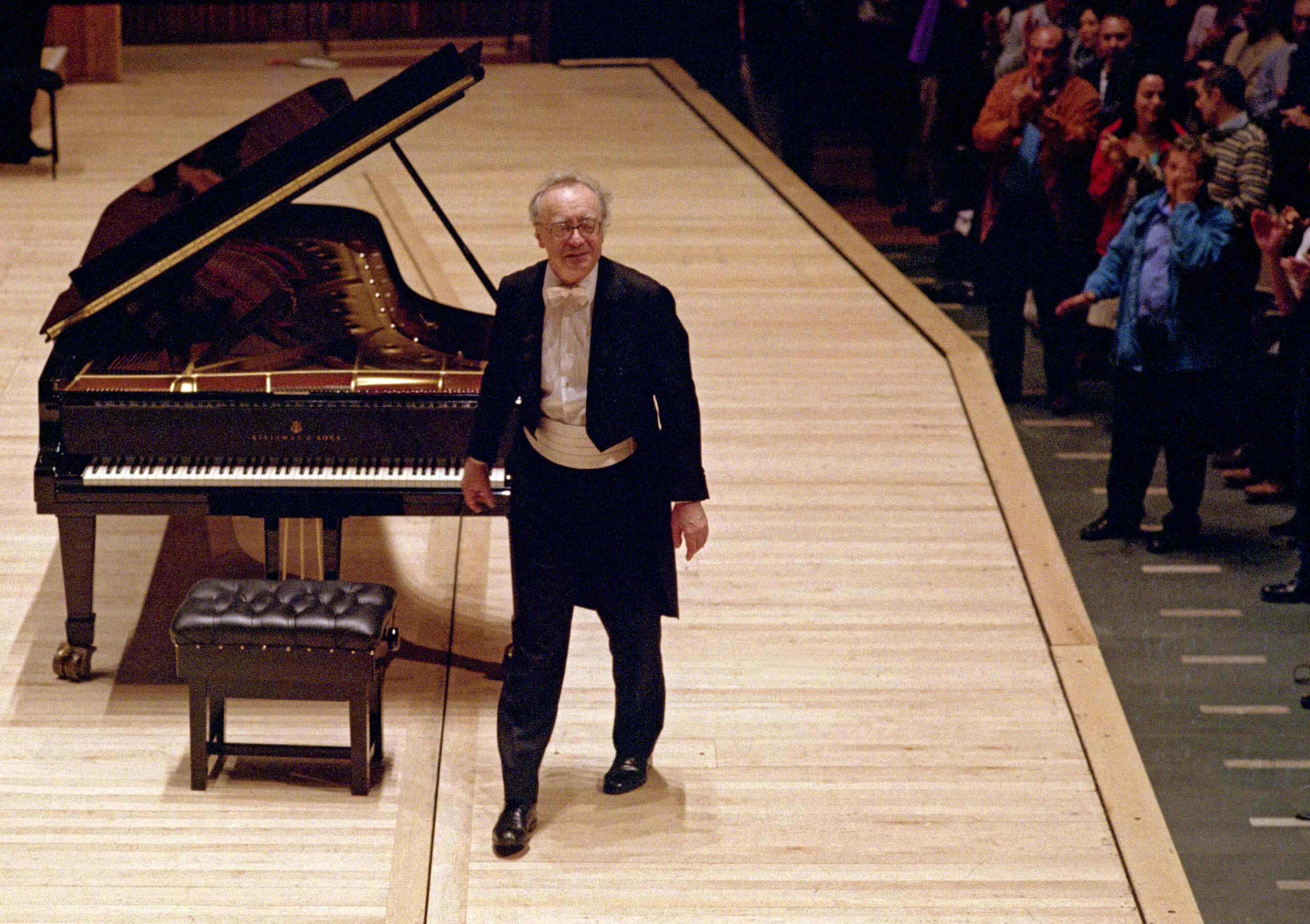 Concert pianist in tailcoat and white tie stands by grand piano as audience stands in applause