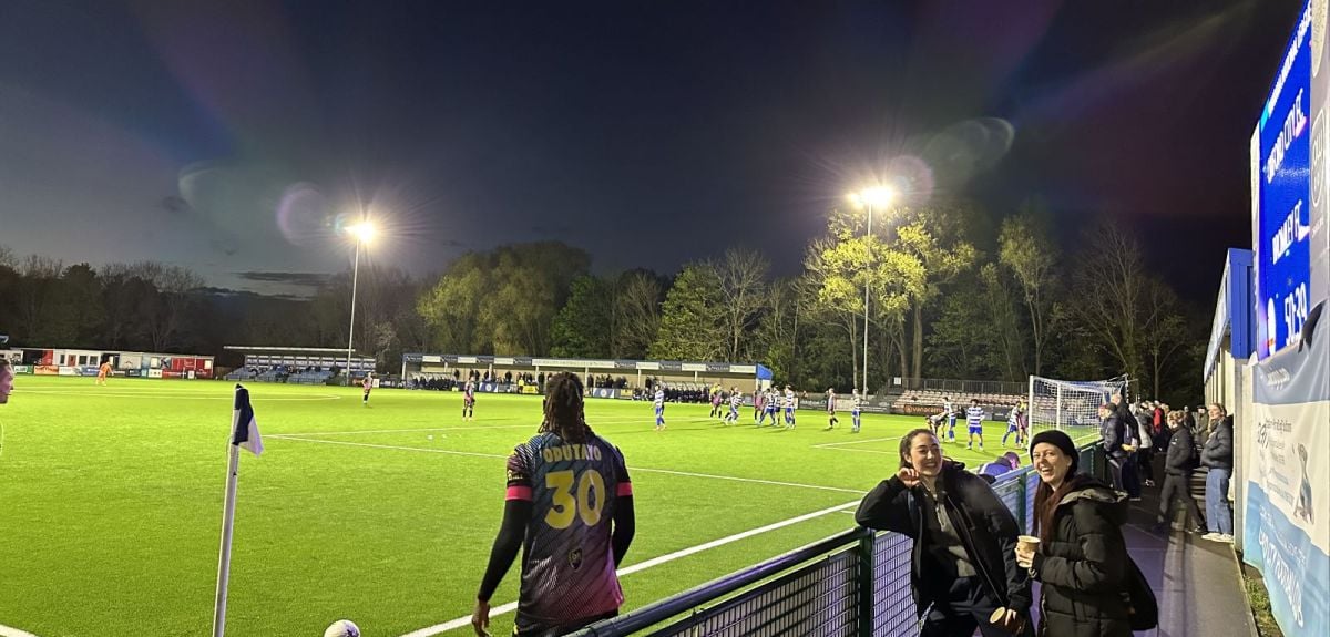 Evening football match under floodlights at a community ground, with players on the pitch, spectators in the stands, and two people smiling at the camera by the pitchside barrier.