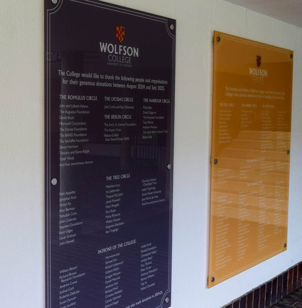 Photograph of two donor boards mounted on a white wall at Wolfson College, Oxford. The board in the foreground, printed in dark blue, lists names of donors recognised for their contributions between August 2024 and July 2025. The Wolfson College crest appears at the top. A second amber-coloured donor board is visible in the background.
