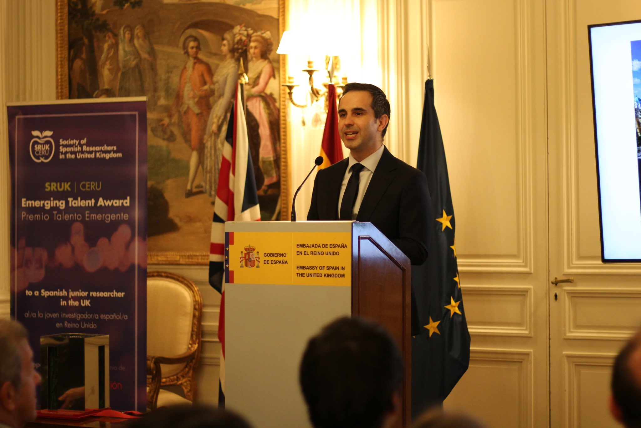 Jesus Lizard stands at a lectern and speaks in front of GB and EU flags