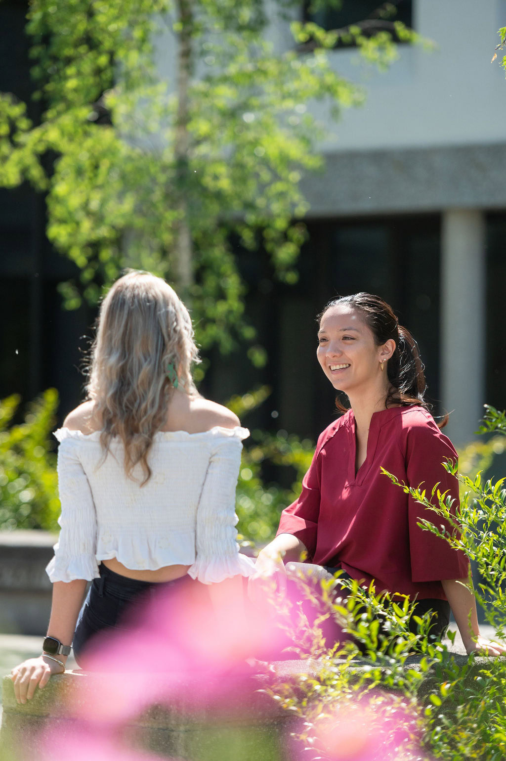 Two females sit in conversation on a bench in a garden, one facing the camera in a red top, the other, back to camera, in a white top.